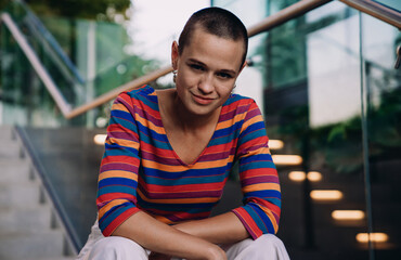 Stylish young adult with short hair sits casually on steps, looking directly at camera with slight...