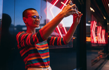 Confident young Asian woman smiling while snapping a selfie in front of vibrant neon arrows,...