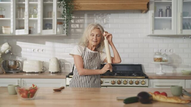 Portrait of mature housewife dancing with rolling-pin in kitchen, good mood. Cheerful grandmother enjoying favorite music and singing alone at home, do housework, cooking and feeling good, have fun