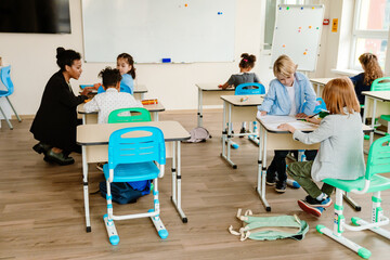 Obraz premium A group of six schoolchildren sit at desks while a teacher talks and sits next to one of them, at school, in a classroom