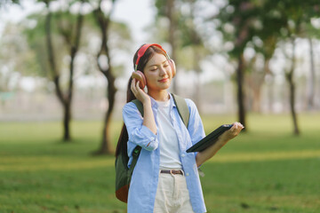 Smiling young woman with headphones and tablet, enjoying music outdoors in a sunny park, surrounded...