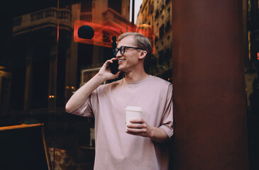 Casual young man standing outside at night, smiling during phone call, holding coffee cup, neon...