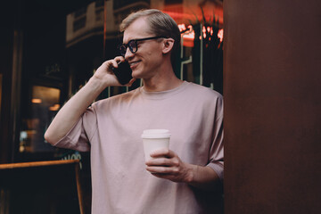 Relaxed young professional enjoying casual phone call with coffee to go, standing outside a café illuminated by colorful neon lights, reflecting modern freelancer lifestyle and urban energy