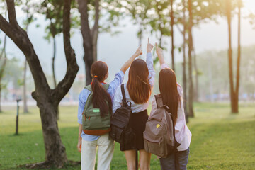 Fototapeta premium Group of students with backpacks joyfully pointing their index fingers up in a park, basking in the warm glow of a sunset