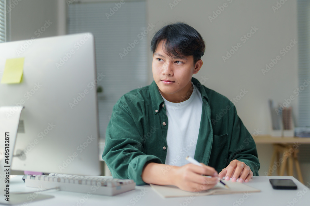 Poster concentrating young asian man taking notes in a notebook while working on a computer in a bright, co