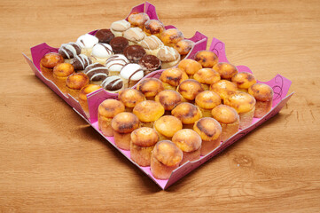 A tray of assorted pastries is on a wooden table. The tray contains a variety of cakes, including...