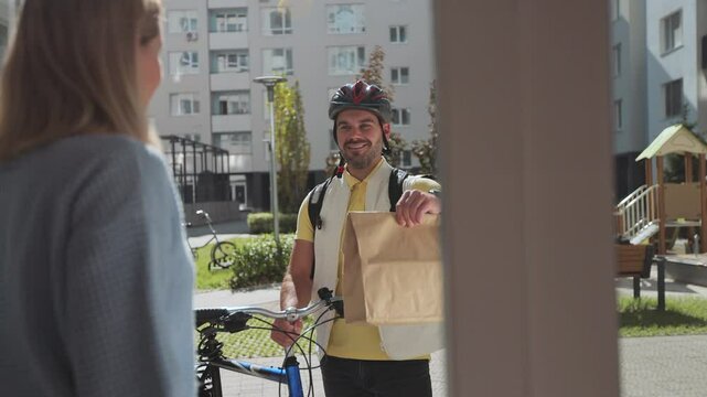 Woman open door to a food delivery man on a bike wearing yellow thermal backpack and recieves order or package with food. Food delivery concept.