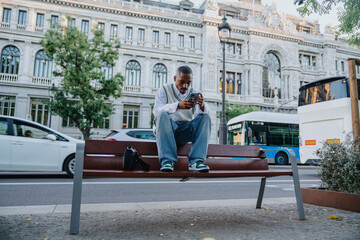 young Black man in stylish urban wear sits on city bench, deeply focused on smartphone. elegant...