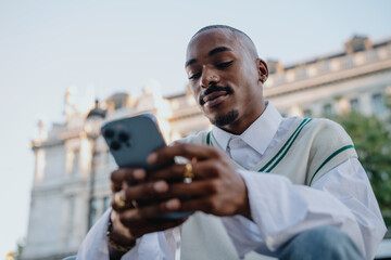 focused Black man in a preppy outfit sits outdoors, scrolling on his smartphone. His stylish look, rings, and well-groomed appearance reflect a modern, sophisticated urban lifestyle