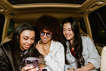 Plakat A group of three female friends sit and laugh while looking at a phone that one of them is holding, in a car