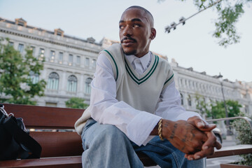 A young Black man in a trendy vest and white shirt sits on a bench, gazing into the distance. His...