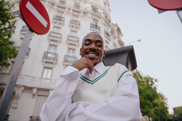 charismatic Black male poses in stylish outfit, resting chin on his hand while smiling. wide-angle...