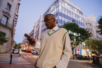 young Black man in a stylish white vest and glasses walks through a modern city at night, checking his smartphone. The illuminated buildings and streetlights create a vibrant urban atmosphere