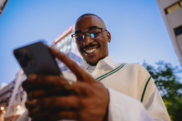 close-up of a young Black happy man in glasses looking at smartphone app with bright smile,...