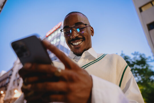 close-up of a young Black happy man in glasses looking at smartphone app with bright smile, reflecting city lights at night. His confident expression and trendy outfit add to modern urban aesthetic