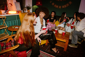 A woman sits and looks at her female friend sitting next to her, they are smiling and holding cups