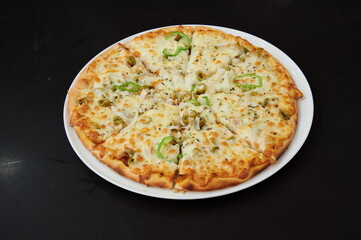 A pizza with cheese and vegetables on a white plate. The pizza is cut into eight slices. The plate is on a black table