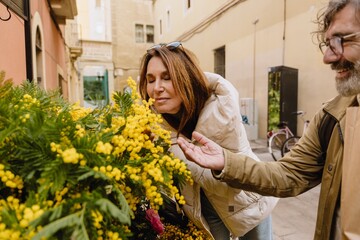 A woman stands with her eyes closed and smells flowers while a smiling man stands next to her, outdoors