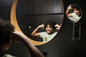 Young boy happily flexing muscles while standing in front of bathroom mirror. Natural light illuminating space, emphasizing his cheerful expression, and striped shirt