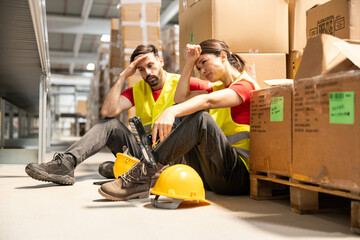Two exhausted warehouse workers sitting on the floor with their helmets off, reflecting on a long...