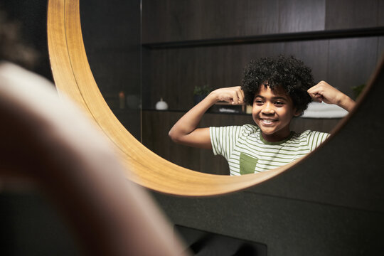 African American boy smiling while flexing his muscles in bathroom mirror, Being proud and confident with curly hair and round mirror