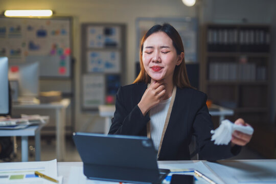 Young Asian businesswoman feeling sick and touching her neck while working late at night in office using tablet and holding tissue paper