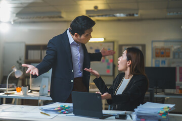 Two Asian businesspeople arguing about work while seated at a desk in an office, facing the stress and pressure of a looming deadline at night