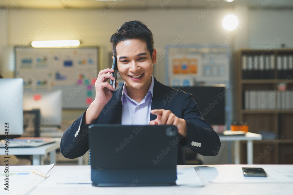 Poster smiling businessman using tablet and talking on mobile phone while working at his desk in modern off