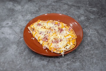 A slice of pizza on a plate. The pizza is topped with cheese and ham. The plate is red and the pizza is on a grey surface