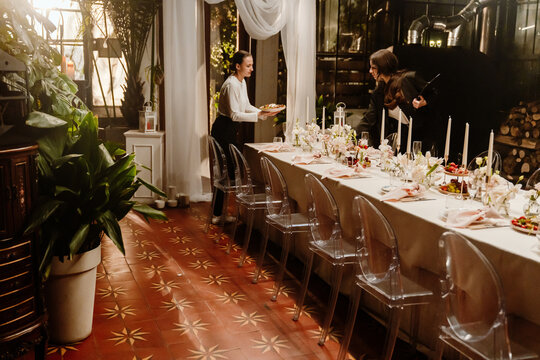 A female waiter carries a plate to the table while a female organizer stands next to her holding a clipboard