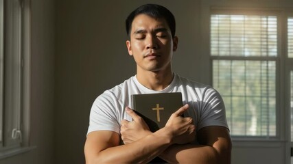 Asian man holding bible with cross in prayer, representing faith. Spirituality and religion footage for religious publication, christian advertising