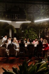A female waiter stands and holds a bottle while a group of ten guests, the bride and groom, sit at...