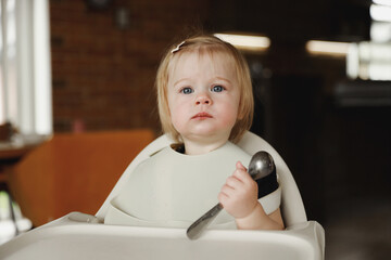Infant girl or toddler sitting on high chair. Eating food by herself using spoon
