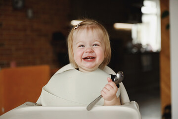 Infant girl or toddler sitting on high chair. Eating food by herself using spoon