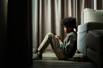 Young boy sitting on floor near window curtains, engrossed in checking phone while leaning back on...