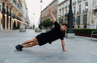 Young Caucasian man practices static side plank on empty city sidewalk, enhancing core engagement,...