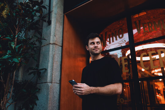 Stylish young man with mobile phone in hand smiling subtly, standing near cozy restaurant entrance with warm lights and greenery, casual evening moment in urban setting.Young male using smartphone app