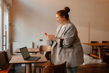 Plakat The female owner stands and smiles while looking at the phone she holds in front of her, in a cafe