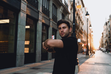Focused young man warming up on urban street during early sunrise. Arm stretch routine shows...