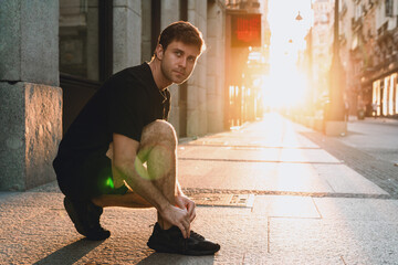 Focused young Caucasian man kneeling to tie his sneakers on city street bathed in warm morning...