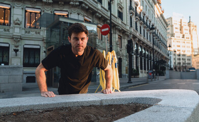 Determined young Caucasian man doing push-ups against concrete bench in early morning city, maintaining intense workout routine surrounded by beautiful European architecture