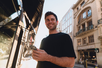 Smiling young man holding smartphone with earbuds in, enjoying mobile phone app in warm city sunlight, surrounded by elegant architecture and street reflections. Enjoying online communication