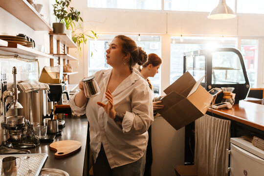 A female owner stands behind the bar and holds a teapot while a female employee next to her holds a box, in a cafe