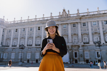 Cheerful young tourist using mobile phone near famous Madrid palace, enjoying solo travel and discovering new places with help of apps, maps, tickets and digital tools on vacation