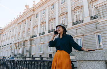 Fototapeta premium Smiling young Asian woman in hat and glasses talking on smartphone while traveling in Madrid, enjoying summer vacation, roaming service, and cultural cityscape near historic architecture