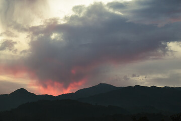 Dark mountain silhouettes beneath dramatic clouds glowing with fiery red and pink hues during a...