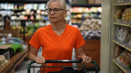 Slow motion front view of pretty mature blonde woman pushing shopping cart through a grocery store and getting plastic bag for produce.