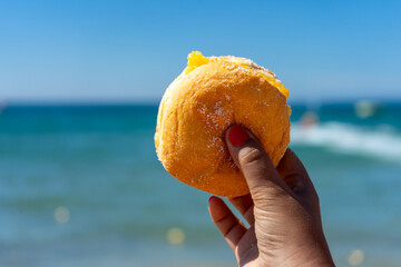 A woman’s hand holding a traditional Portuguese Bola de Berlim against the vibrant blue sea,...