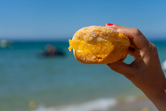 A woman&rsquo;s hand holding a traditional Portuguese Bola de Berlim against the vibrant blue sea, capturing a perfect summer beach moment.