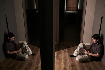 Teenager sitting on wooden floor using smartphone in dimly lit hallway with dark walls and closed...